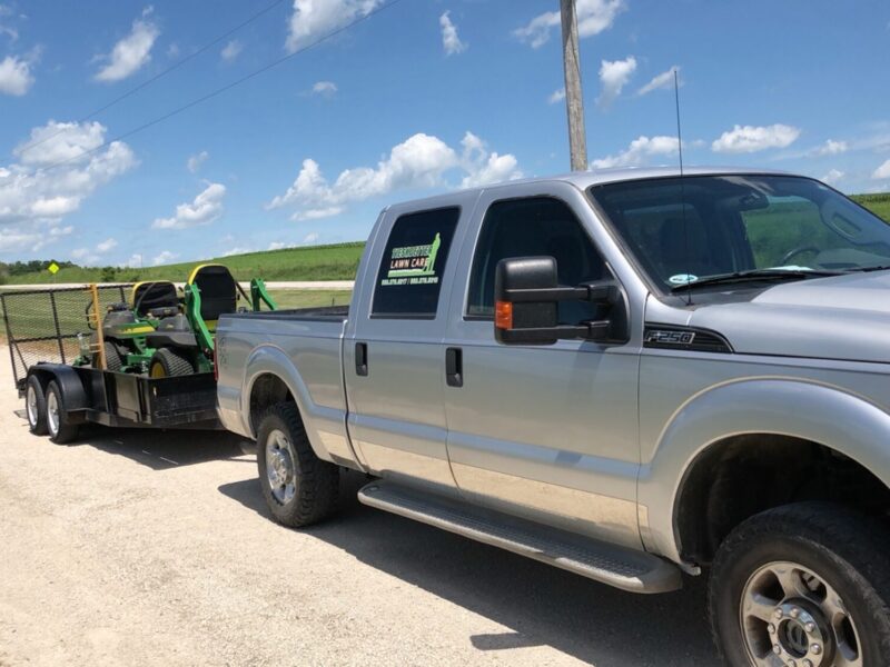 A silver truck towing a trailer with green equipment on a rural road under a blue sky with scattered clouds.