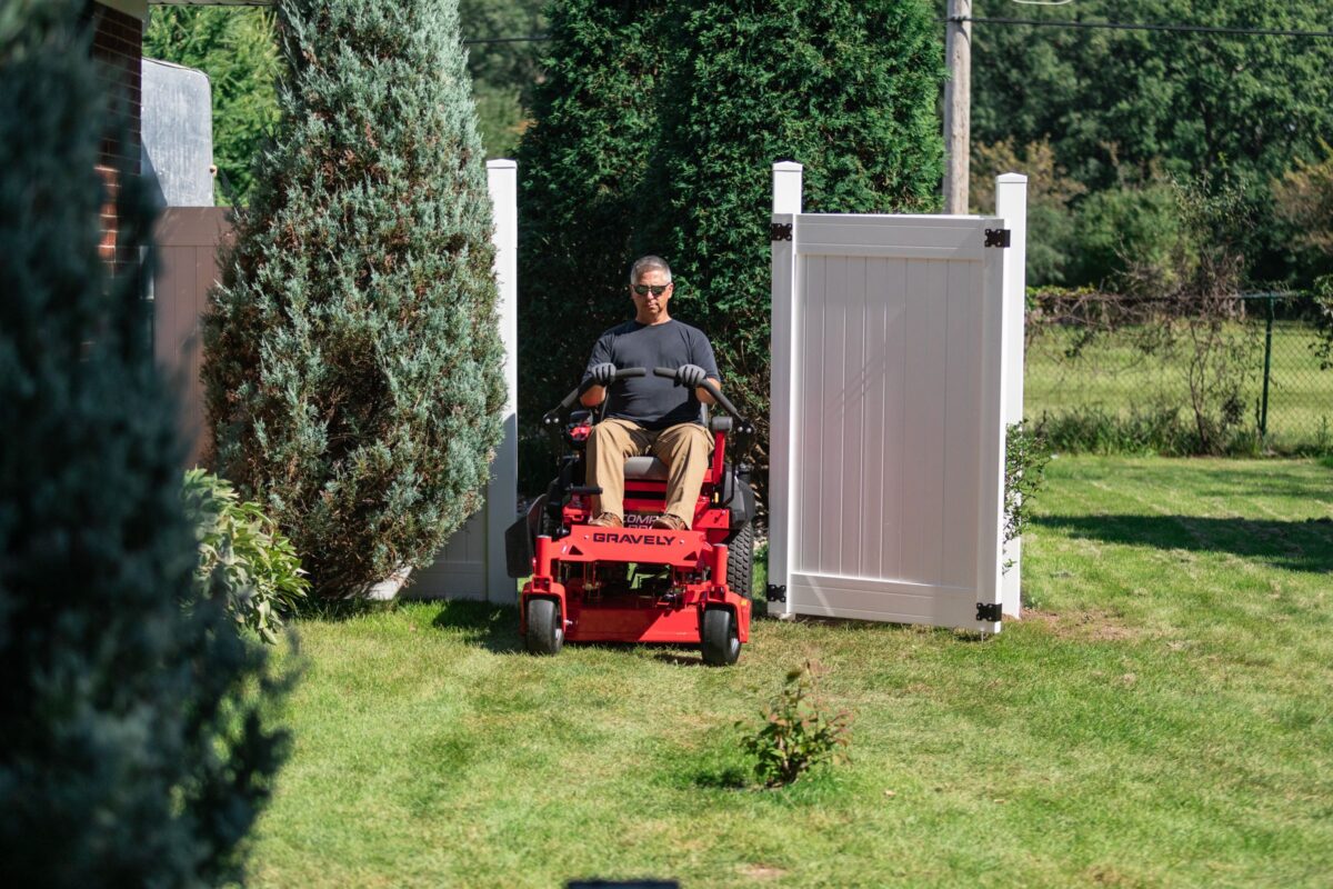 Person mowing the lawn on a red riding lawn mower, exiting through a white wooden gate, surrounded by green trees and grass.