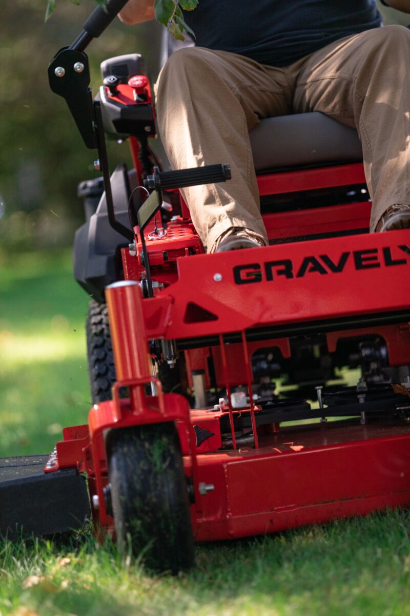 Person operating a red Gravely lawn mower on grass, focusing on the front wheel and cutting mechanism.