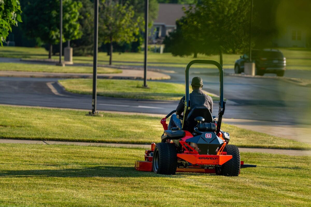 Person mowing grass on a ride-on lawnmower in a park, facing away from the camera, with trees and a road in the background.