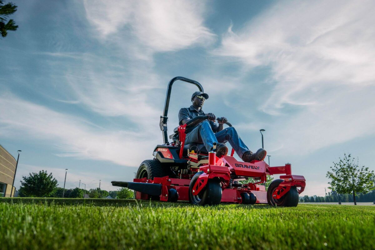 A person operates a red lawn mower on a grassy field under a partly cloudy sky.