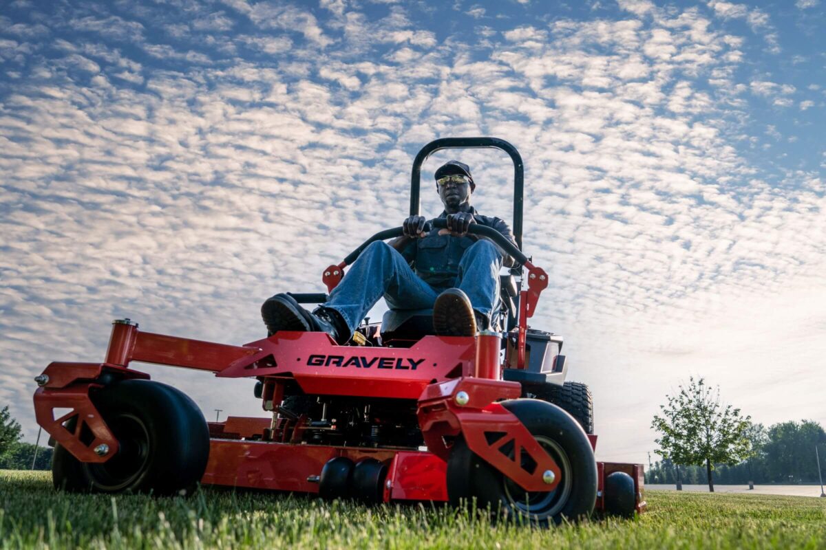 Person operating a red Gravely riding lawn mower on grass under a partly cloudy sky.