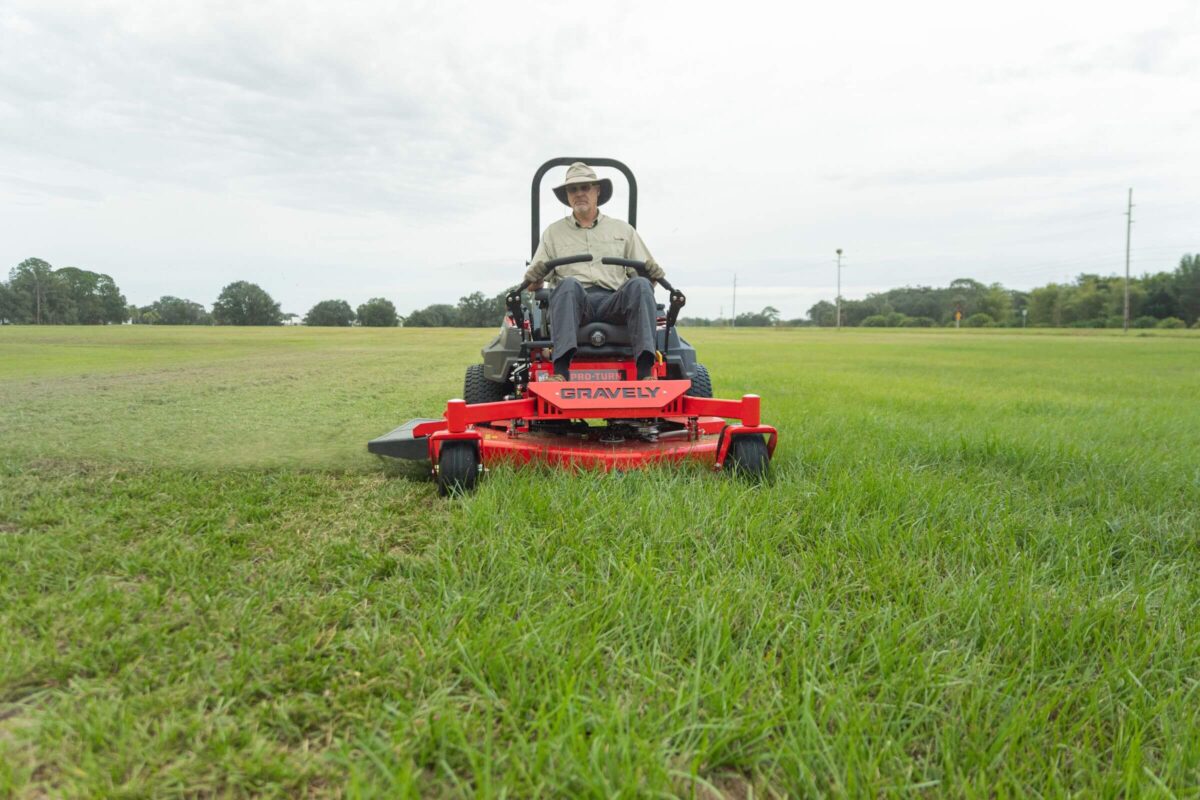 A person wearing a hat drives a red Gravely lawnmower, cutting grass in a large field under a cloudy sky.