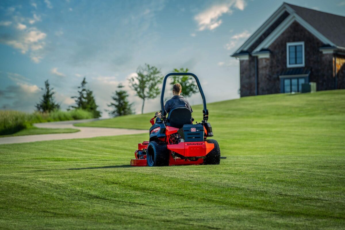 Person operating a red riding lawn mower on a large green lawn with a house and trees in the background.