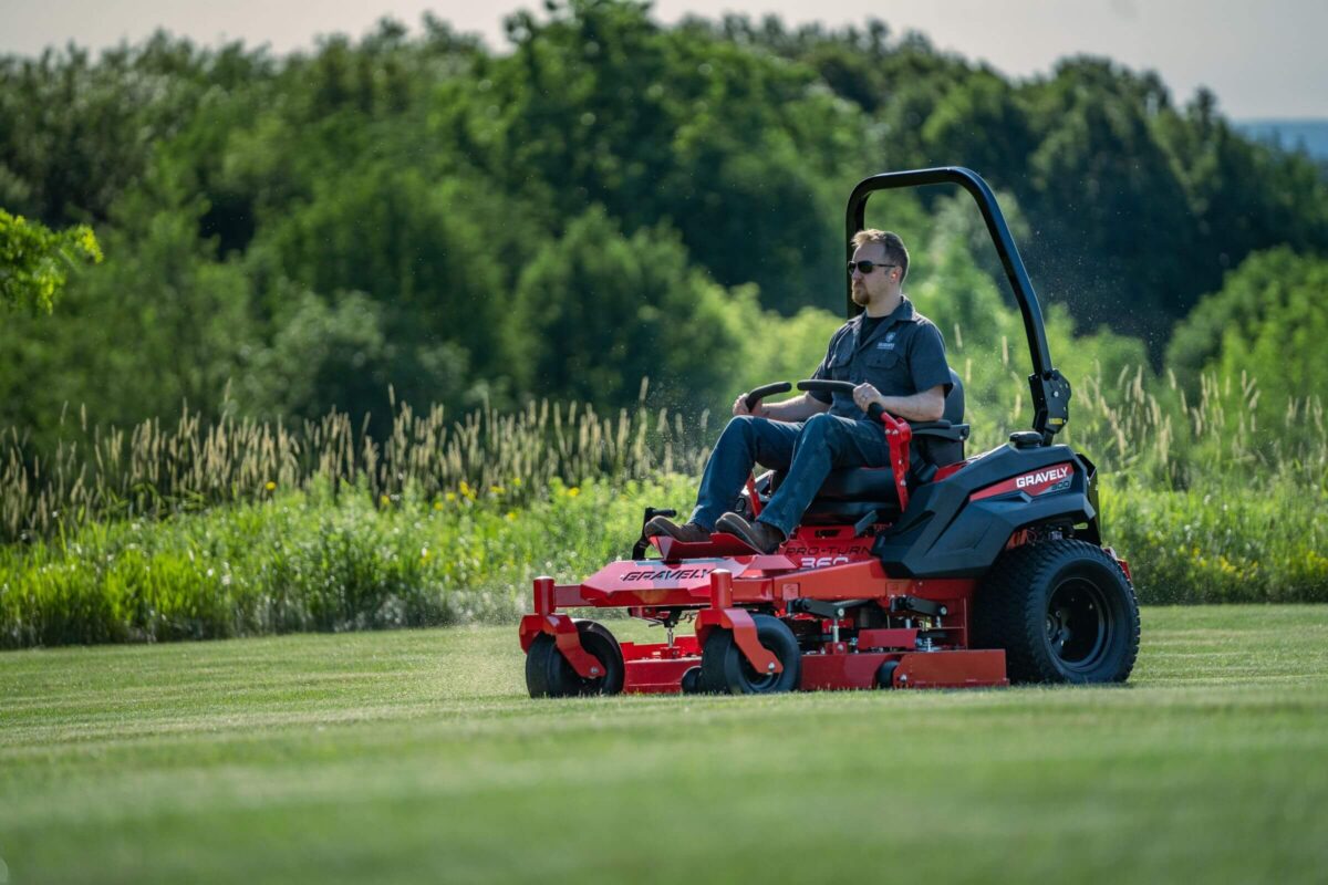 Person mowing grass on a red riding lawn mower in a lush, green field with trees in the background.