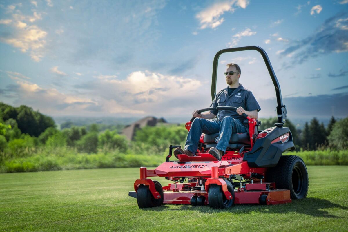 A person operates a red lawn mower on a grassy field under a partly cloudy sky.