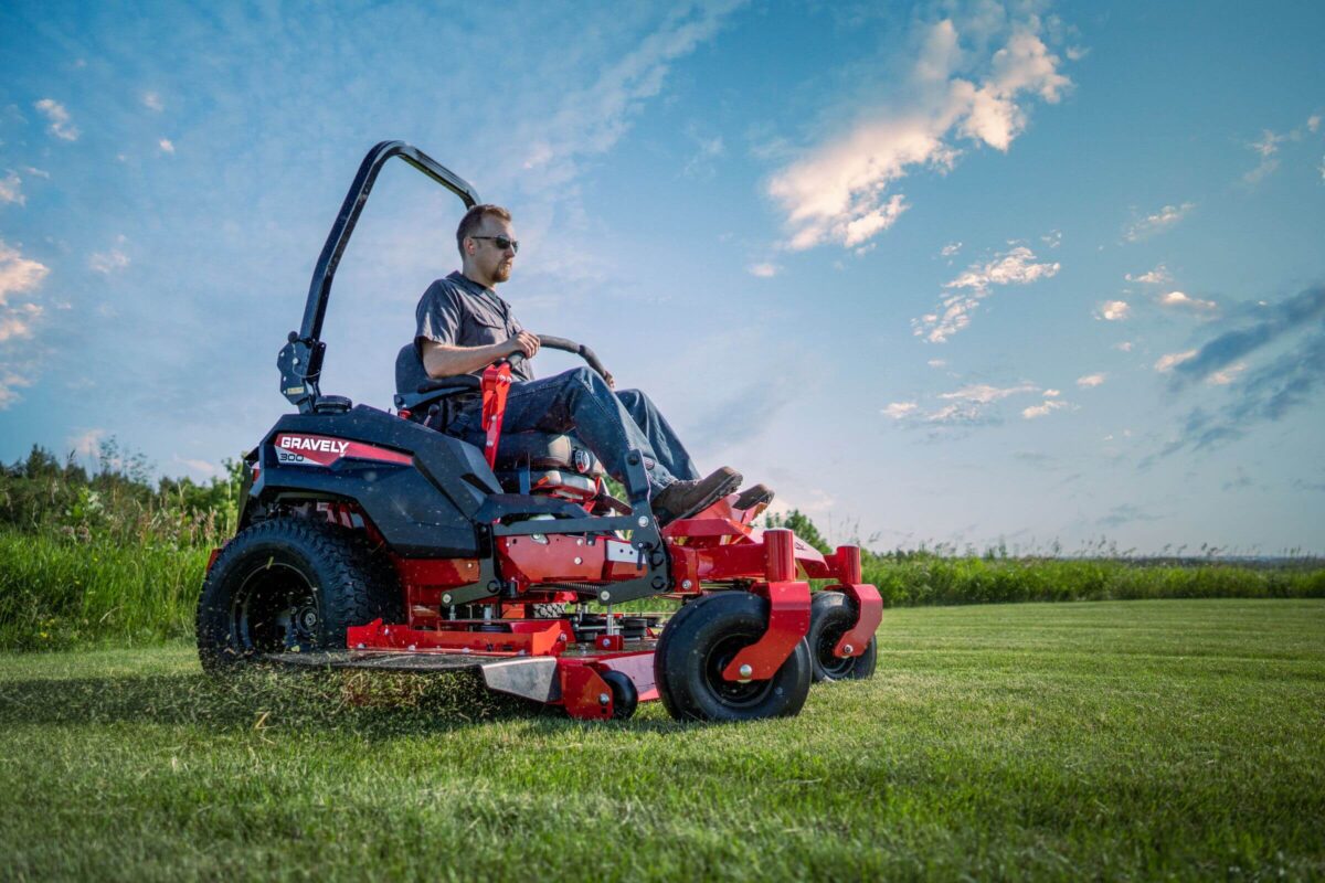 Person mowing grass on a red riding lawn mower in an open field under a blue sky with clouds.