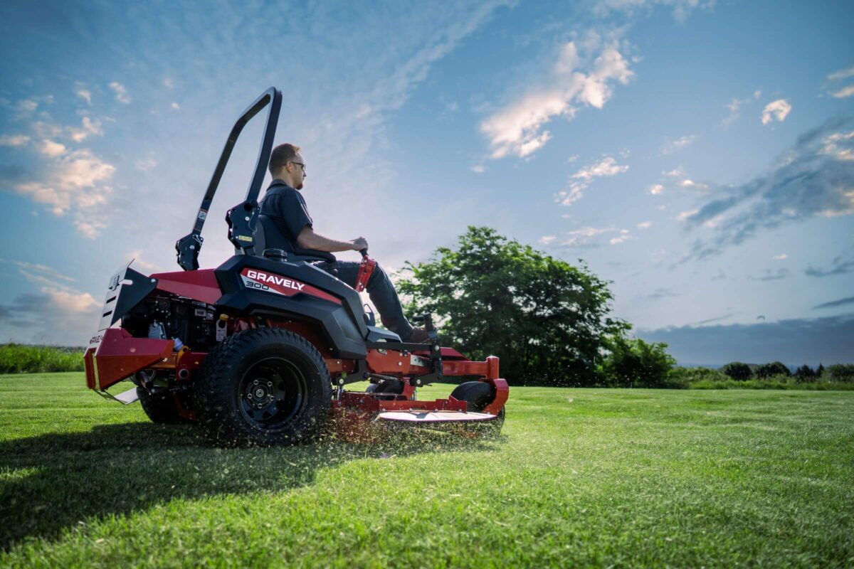 Person riding a red riding lawn mower across a green field with trees in the background under a partly cloudy sky.