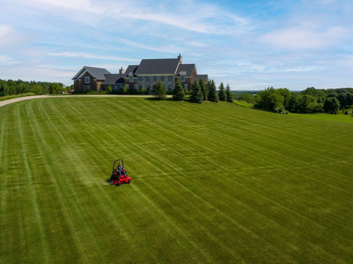 A person on a lawnmower cuts grass on a large, well-maintained lawn in front of a large house. The sky is partly cloudy.