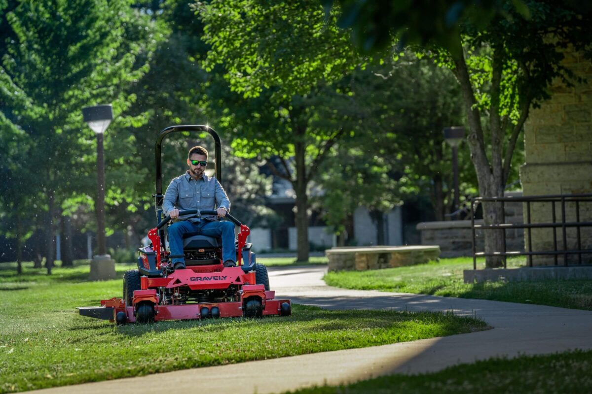 Person driving a red lawnmower on grass near a concrete path, surrounded by green trees and a stone wall.