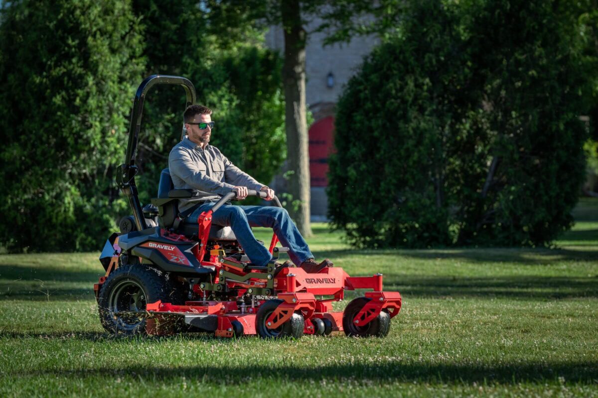 Person driving a red lawn mower on grass, surrounded by trees on a sunny day.
