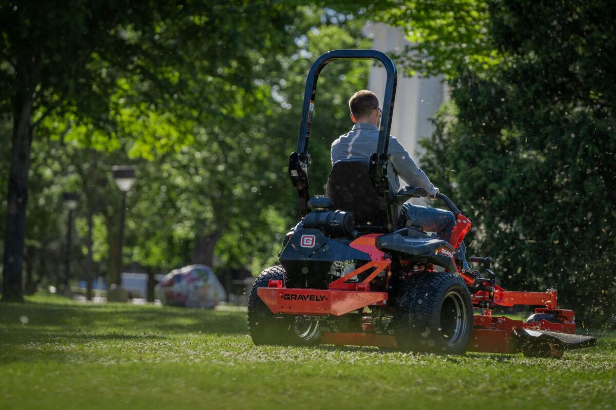 Person mowing grass with a red riding lawn mower in a sunlit park, surrounded by trees.