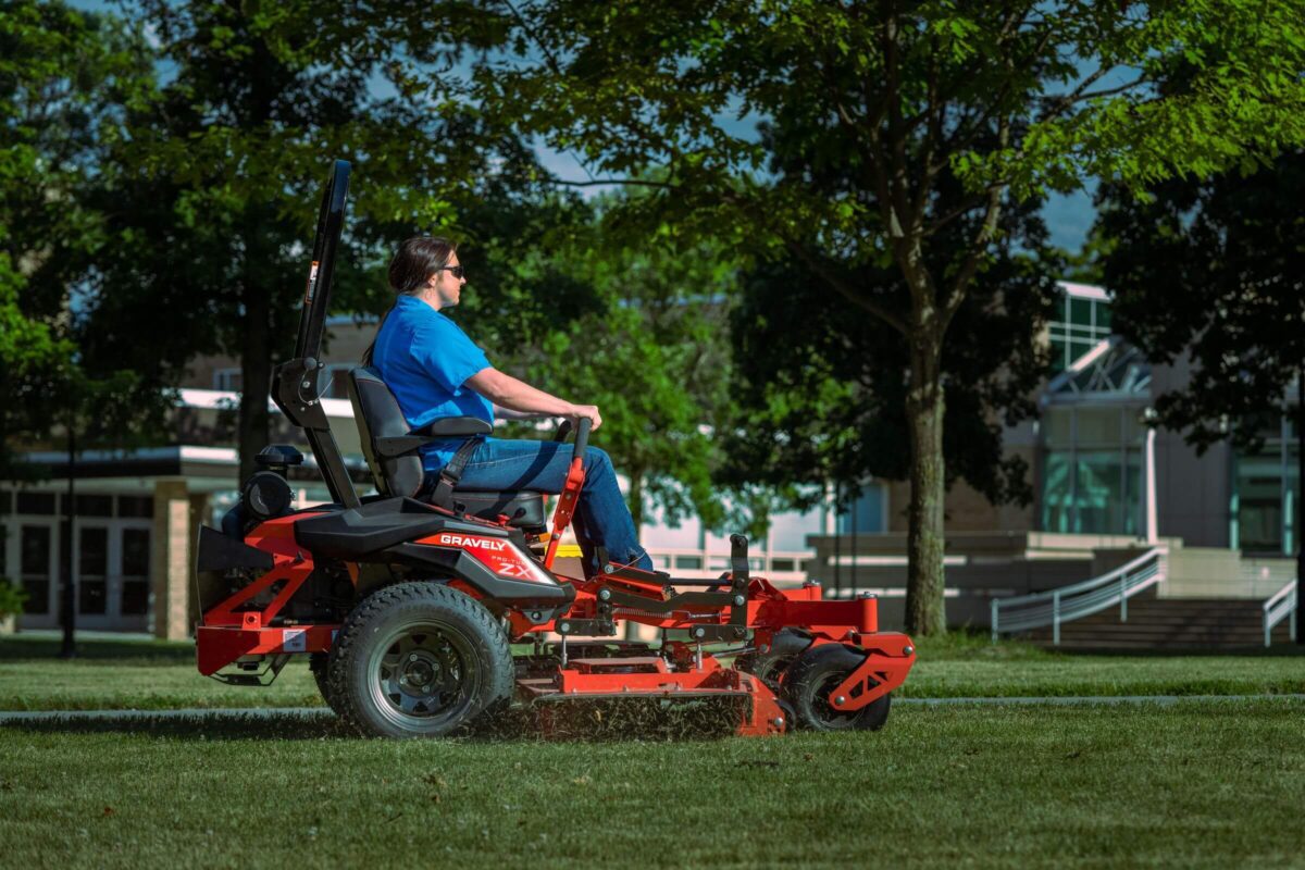 Person mowing grass on a red riding mower in a sunny park with trees and a building in the background.