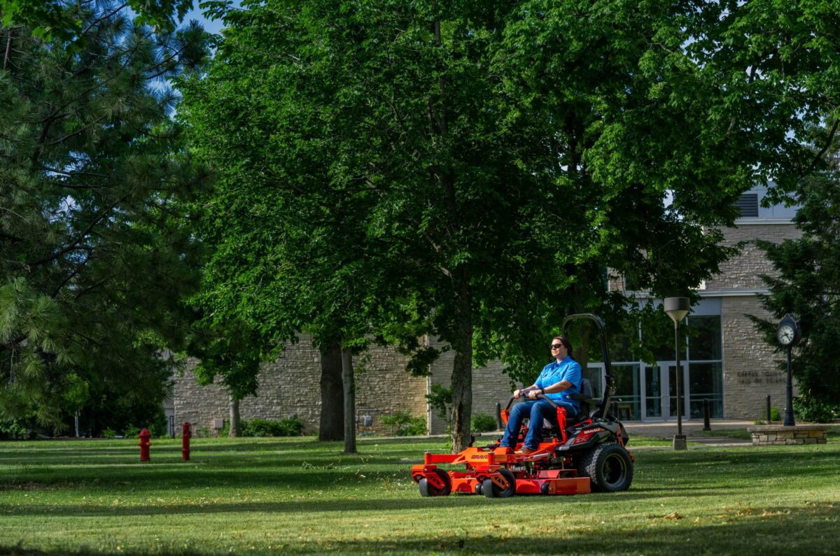 A person in blue operates a red riding lawn mower on a grassy area, surrounded by trees and near a brick building.
