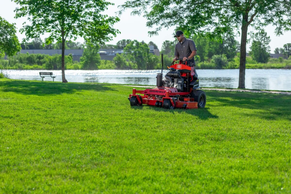 Person operating a red lawnmower on a grassy area near a lake with trees and a bench in the background.