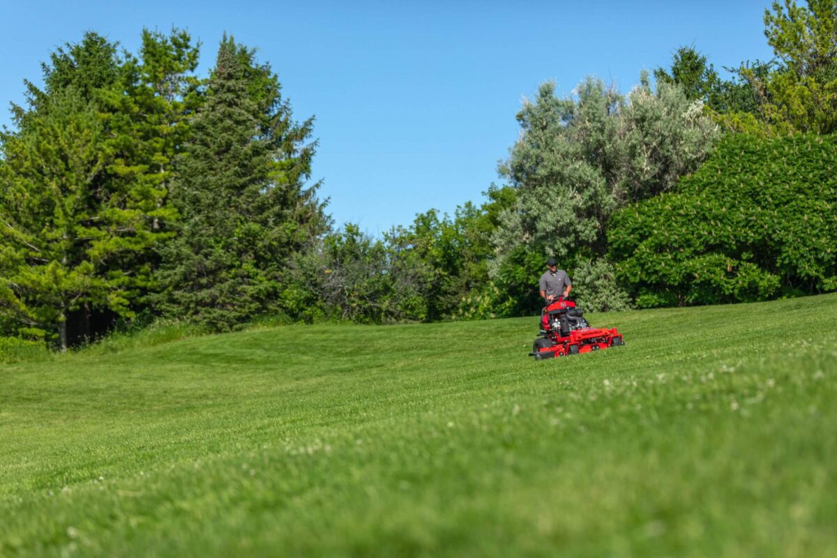 A person operates a red lawnmower on a large grassy field surrounded by trees under a clear blue sky.