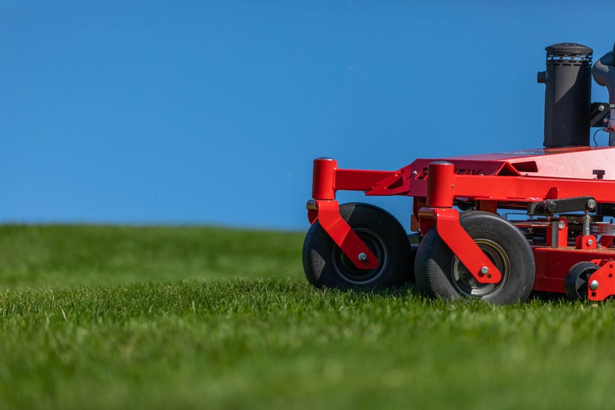 A red lawnmower with large black wheels is shown cutting grass against a clear blue sky backdrop.