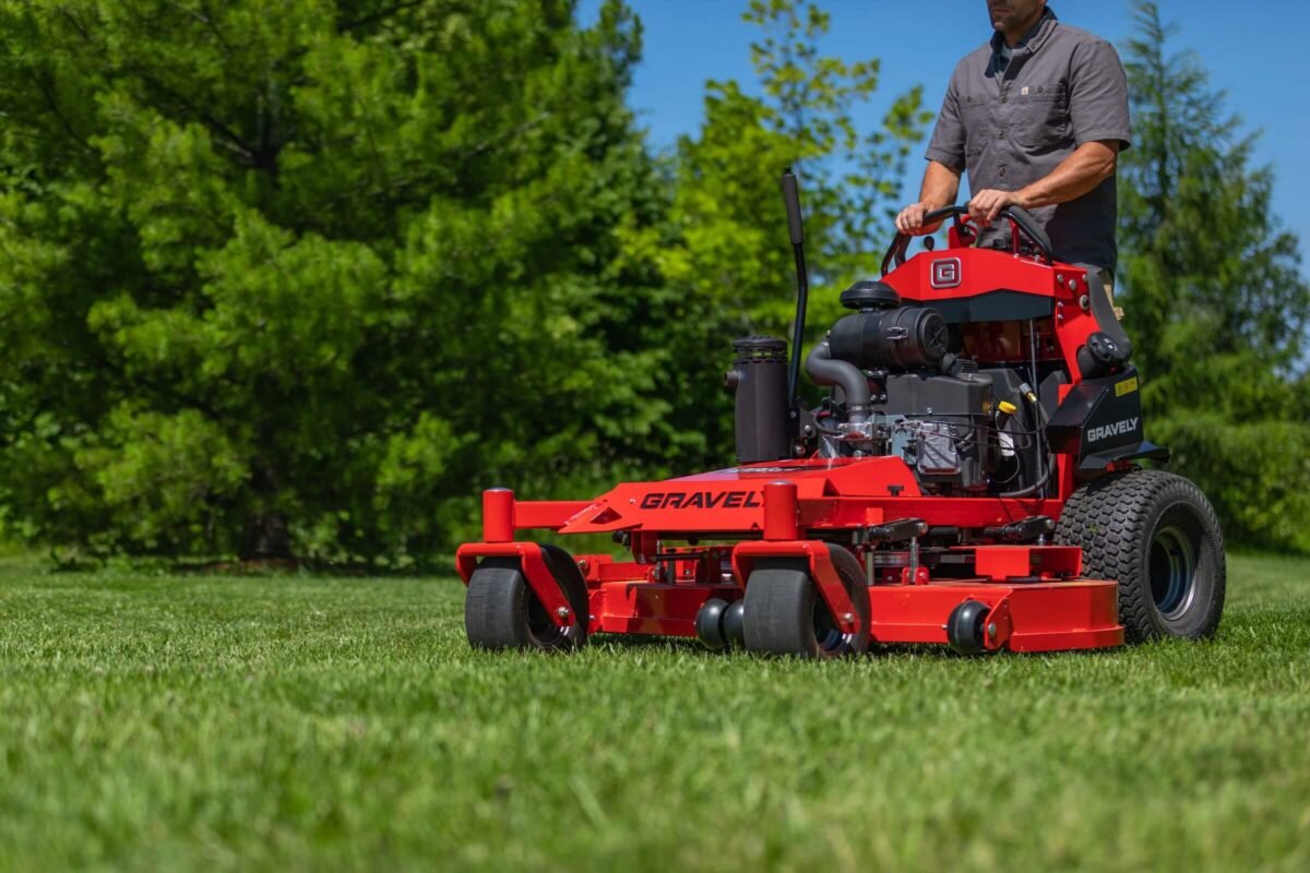 Person operating a red commercial lawn mower on a grassy area with trees in the background.