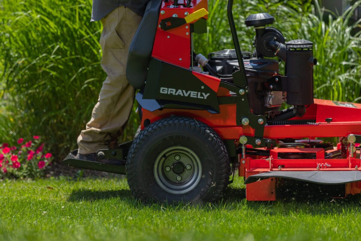 Person operating a Gravely riding lawn mower on lush green grass, with red flowers in the background.