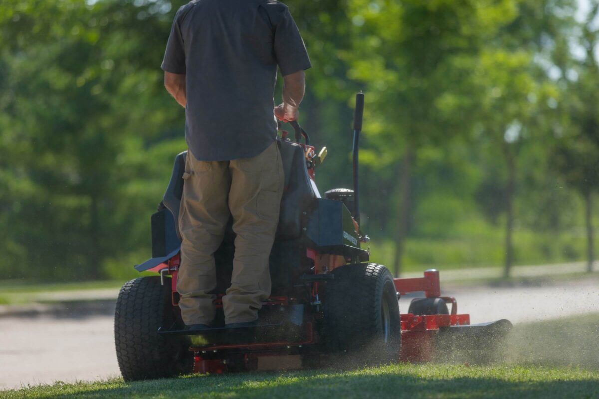 A person operates a red riding lawn mower, cutting grass in a green outdoor setting with trees in the background.