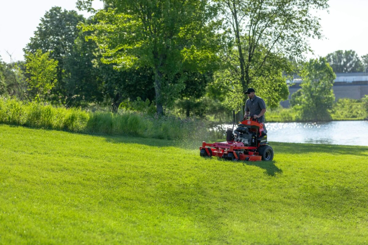 Person mowing a large, green lawn with a red riding lawnmower near trees and a pond on a sunny day.