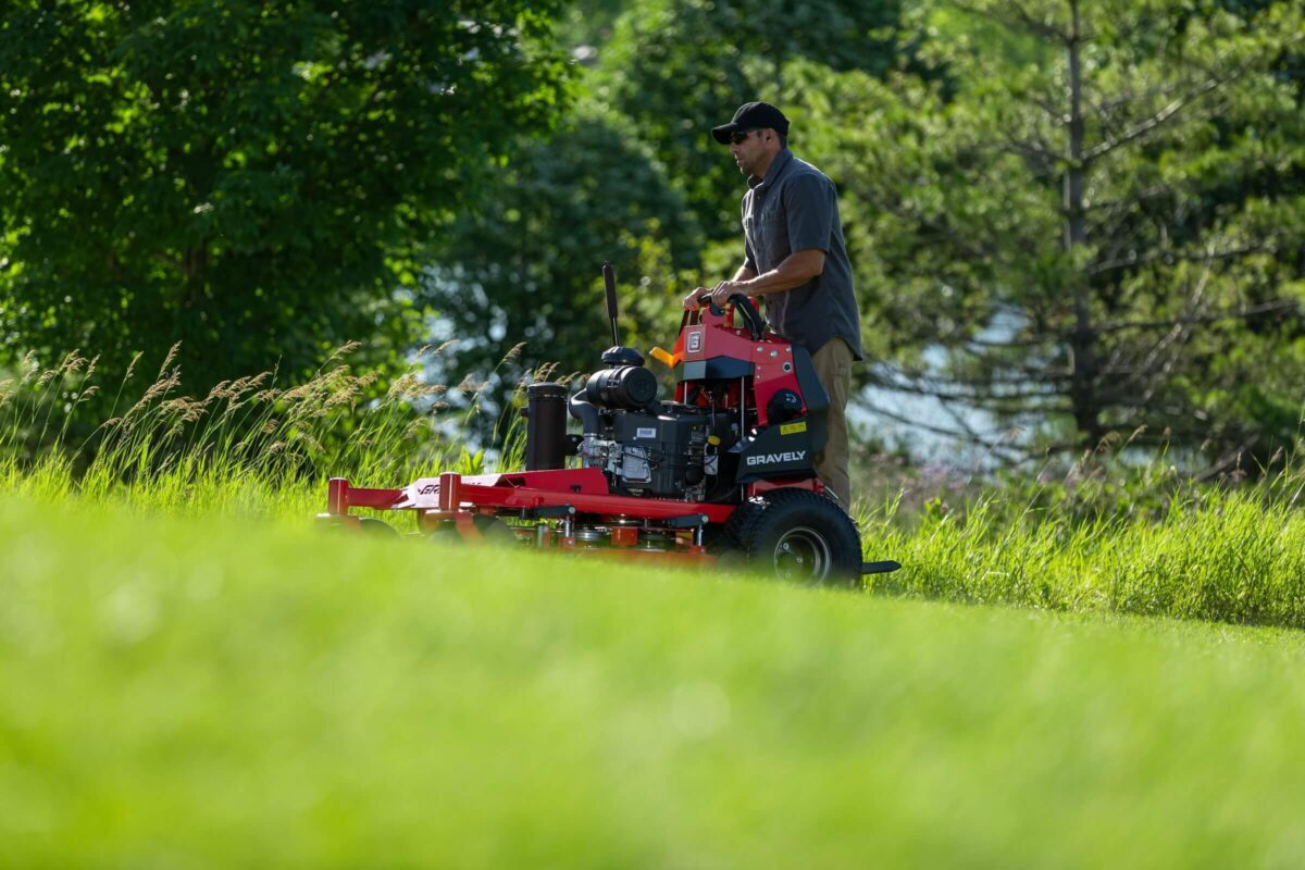 Man operating a red lawn mower on a grassy hill with trees in the background.