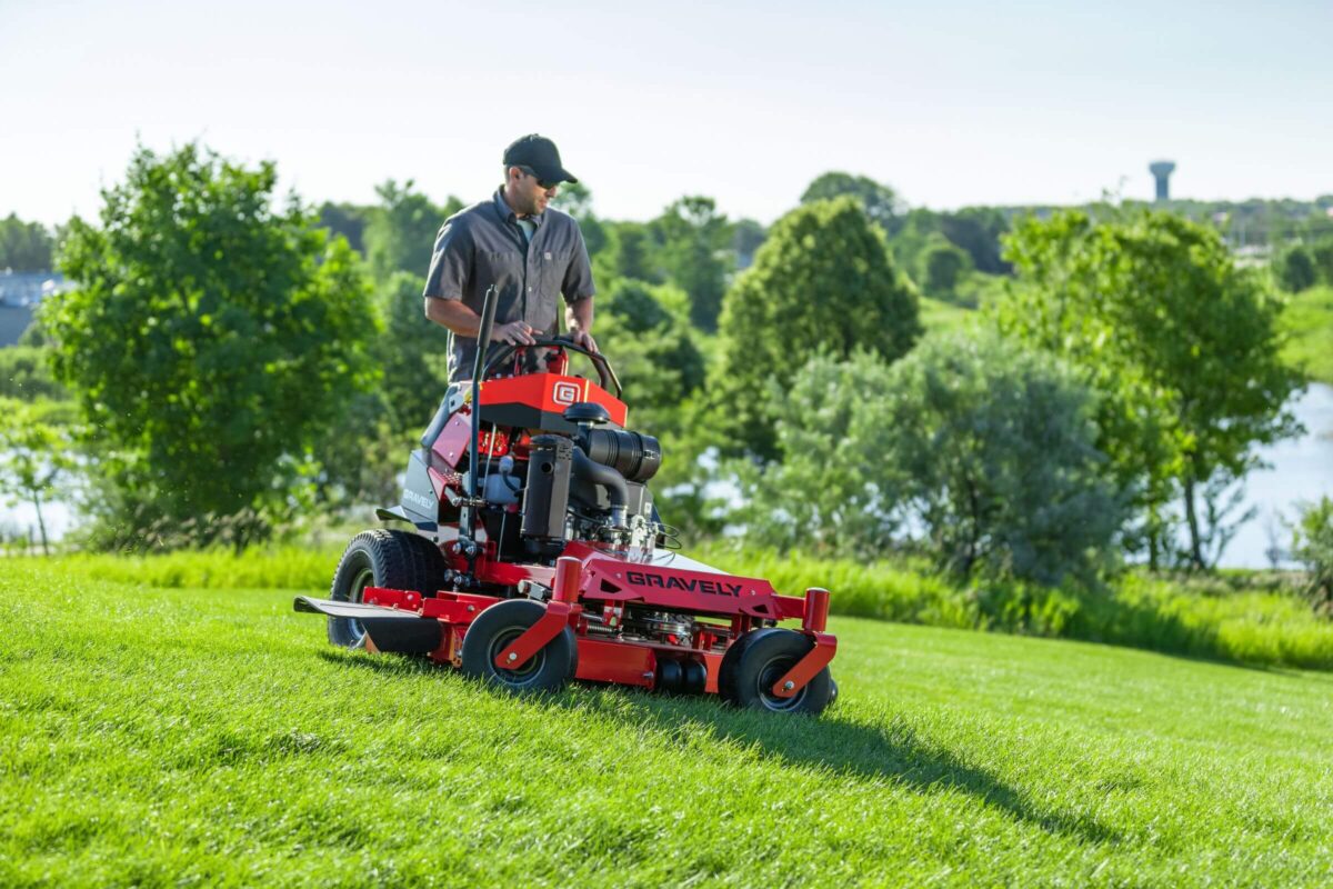 A person operates a red Gravely lawn mower on a grassy slope with trees and a body of water in the background.