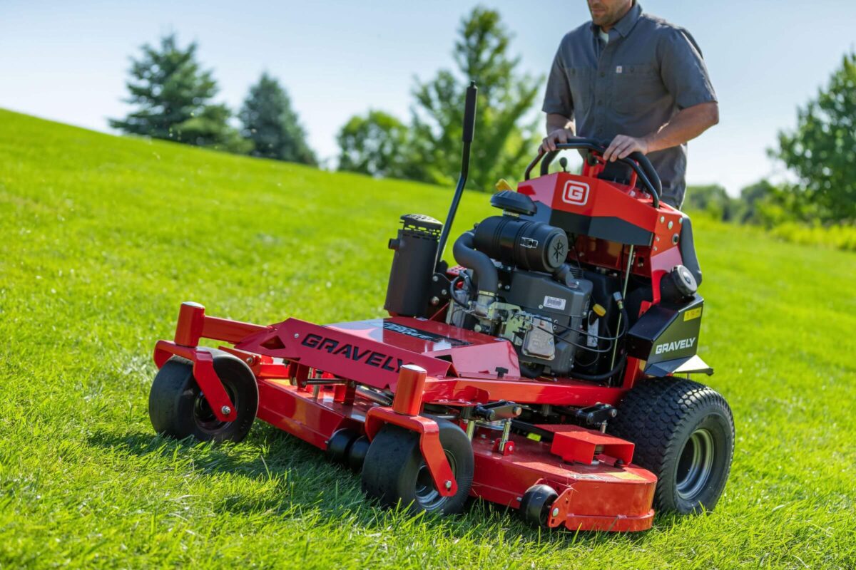 A person operates a red Gravely lawn mower on a grassy hillside under a clear sky.
