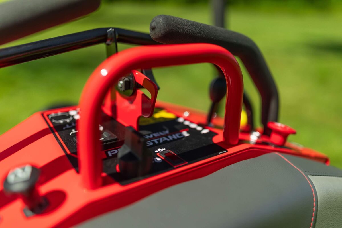 Close-up of the control panel of a red riding lawn mower, showing levers, switches, and a key in the ignition against a blurred green lawn background.