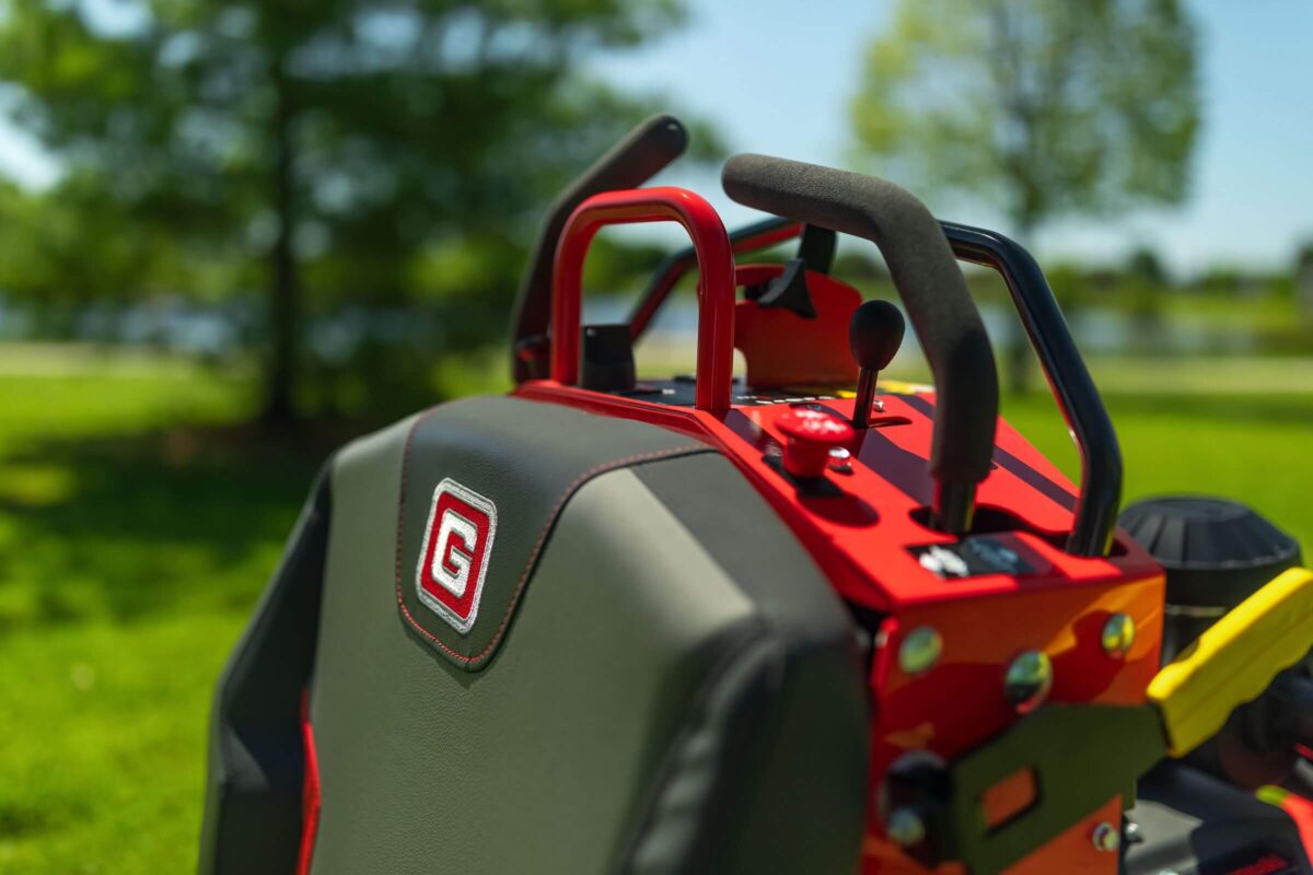 Close-up of a red and black riding lawn mower seat with control levers, set against a background of green grass and trees.