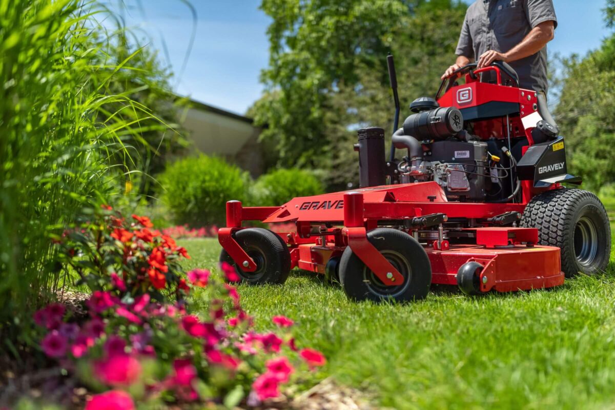 A person operates a red lawnmower on lush grass, surrounded by blooming flowers and greenery in a garden setting.