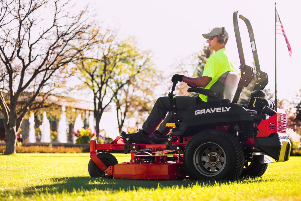 Person in a neon shirt operating a Gravely lawnmower on a sunny day. An American flag and trees are in the background.