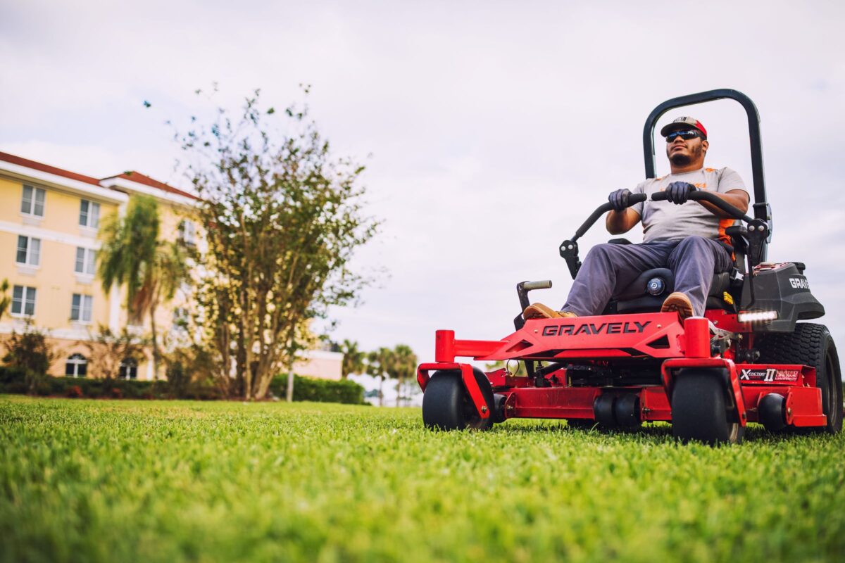 A man operates a red Gravely lawnmower on a grassy area near a tall building. He wears sunglasses, a cap, and gloves, focusing on the task. A large tree is in the background.