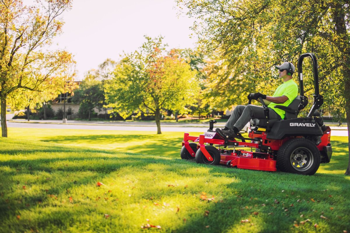 Person operating a red riding lawn mower on a green lawn, surrounded by trees under a clear sky.