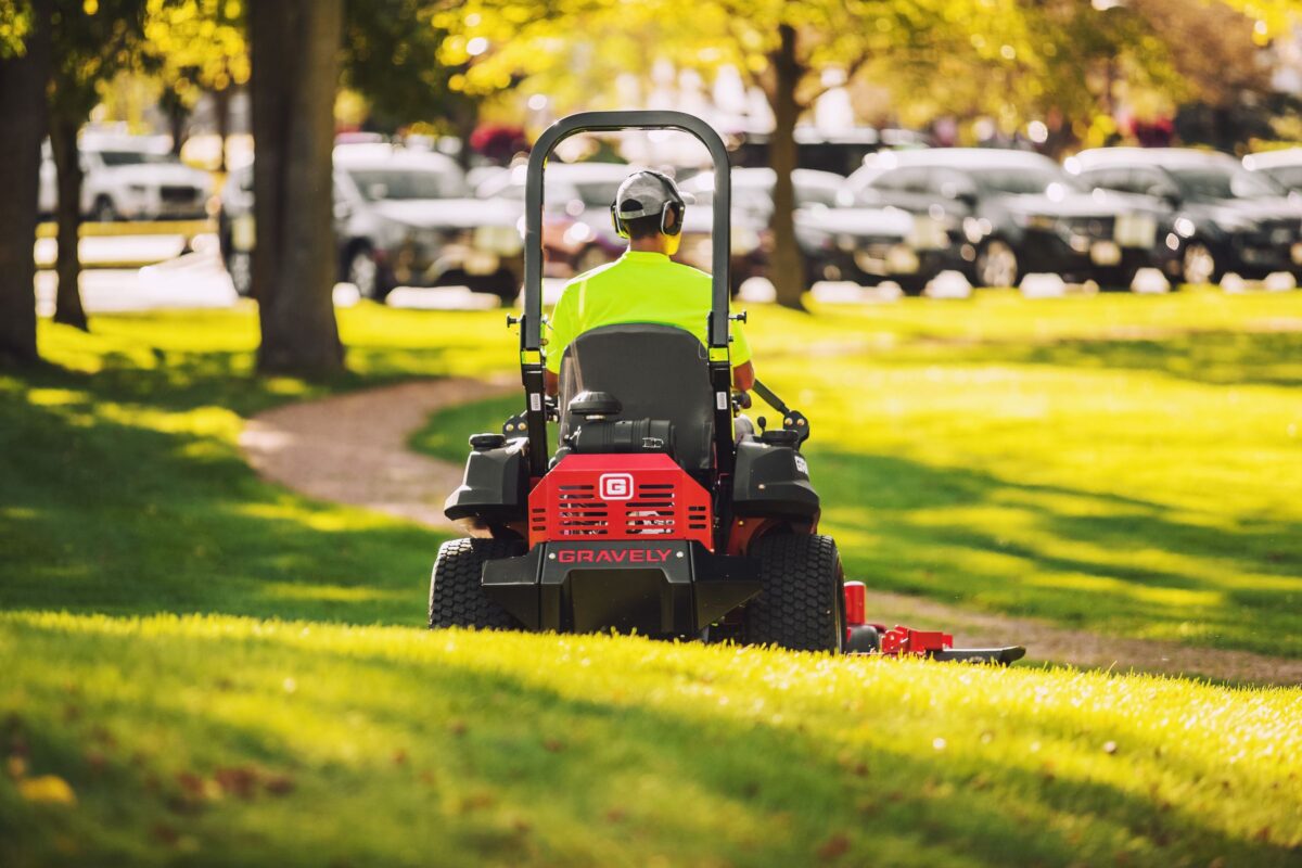 Person operating a red Gravely lawn mower, wearing a neon yellow shirt and headphones, mowing grass in a park with trees and parked cars in the background.