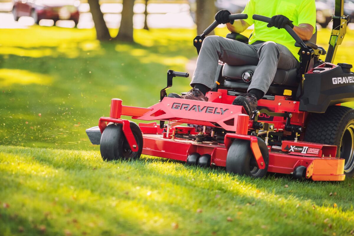 Person operating a red Gravely lawn mower on a grassy lawn, wearing a bright yellow shirt and gray pants.