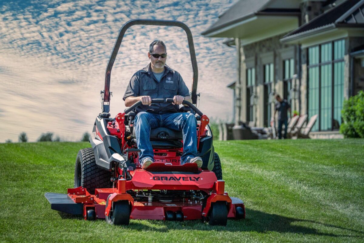 Person operating a red Gravely riding lawn mower on a grassy lawn with a modern house and cloudy sky in the background.