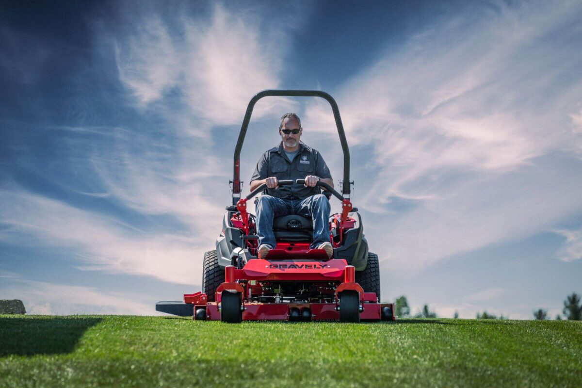 A person rides a red Gravely lawn mower on a well-manicured field under a blue sky with wispy clouds.
