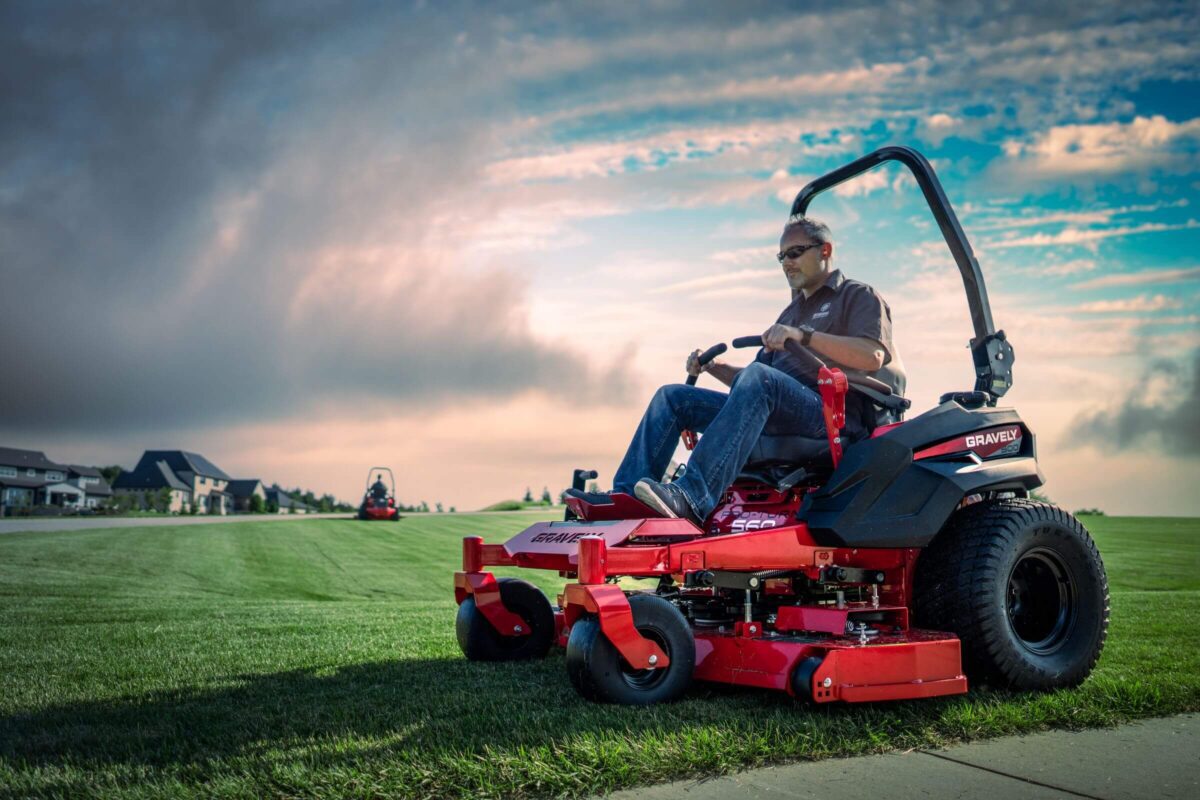 A person operates a red ride-on lawnmower on a large, well-manicured lawn under a cloudy sky. Another lawnmower is visible in the distance.