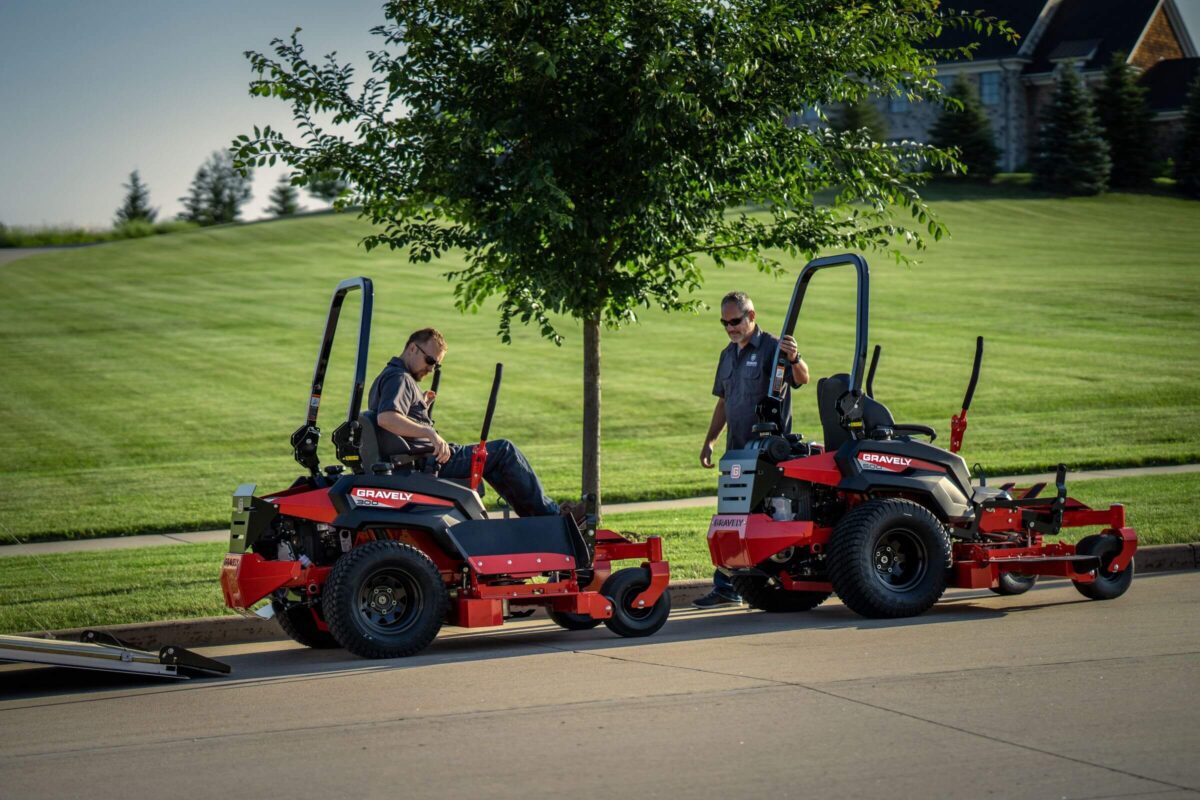 Two people stand near two red lawn mowers on a paved surface. One person is seated on a mower, while the other stands beside it. There is a tree and a grassy area in the background.