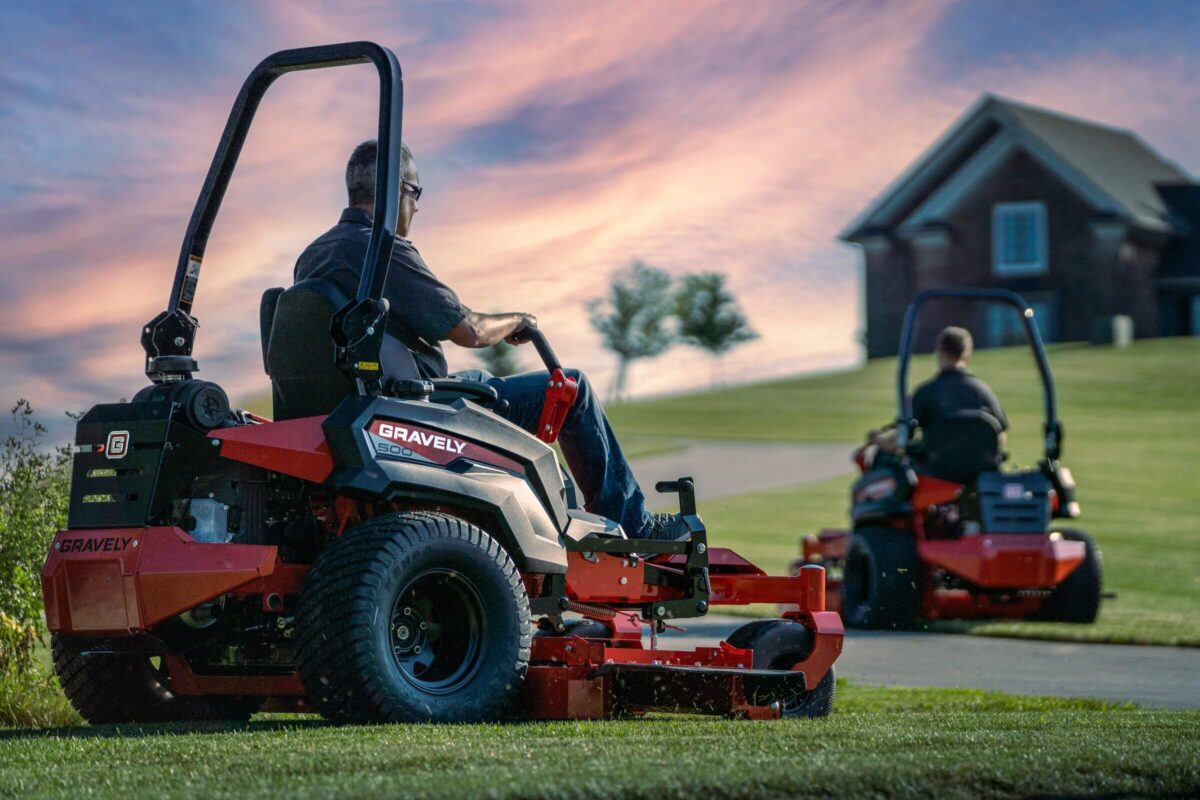 Two people operate red Gravely mowers on a grassy lawn at sunset, with a house and trees in the background.