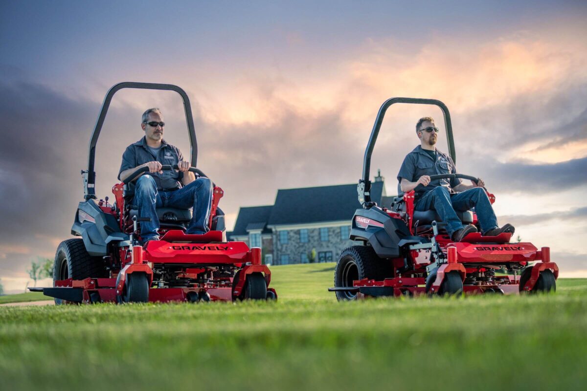 Two people operate red riding lawn mowers on a grassy field with a building and cloudy sky in the background.