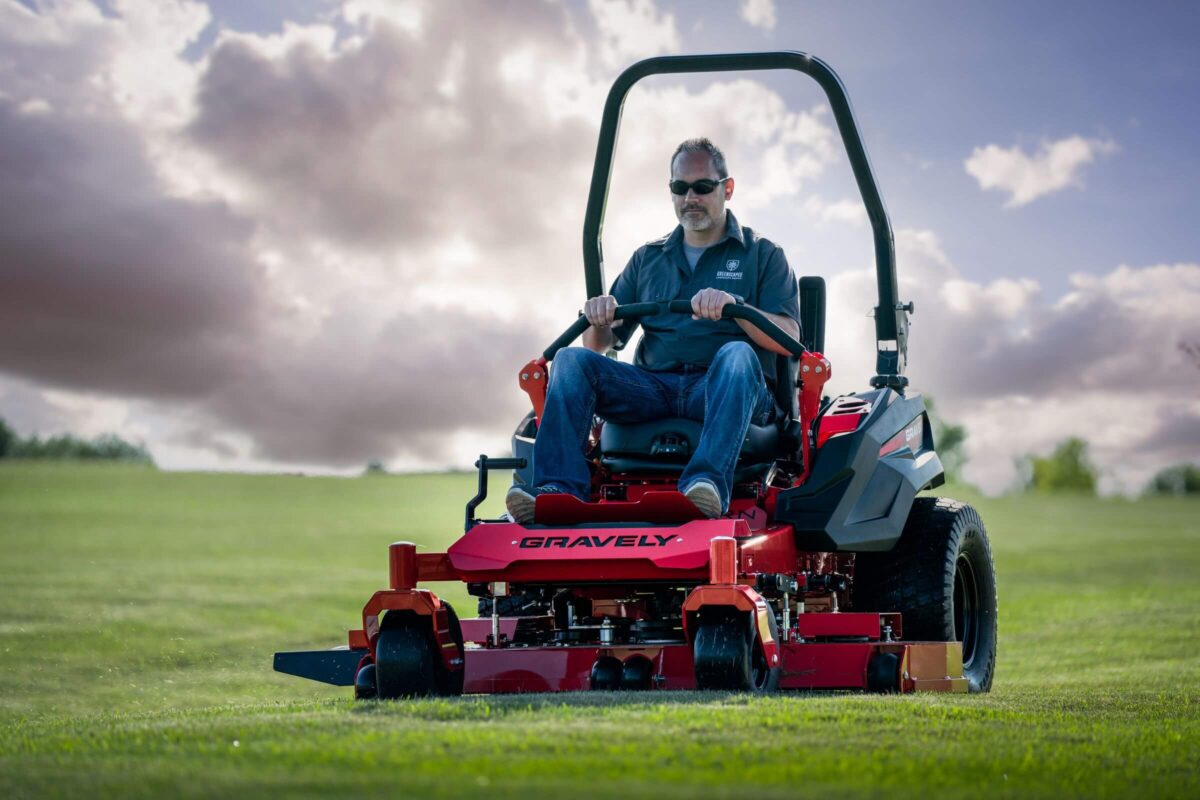 Man operating a red Gravely riding lawn mower on a grassy field under a cloudy sky.