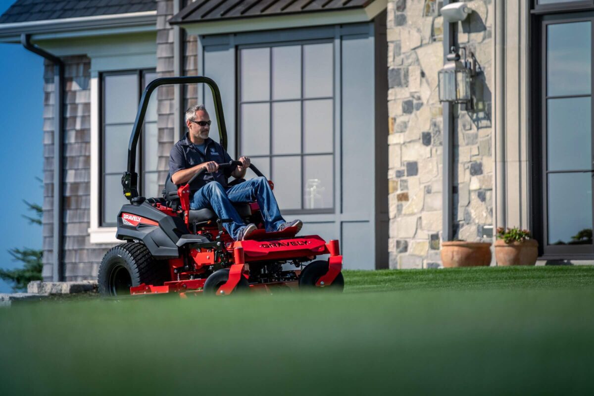 A person operates a riding lawn mower on a well-maintained lawn in front of a house with large windows and stone facade.