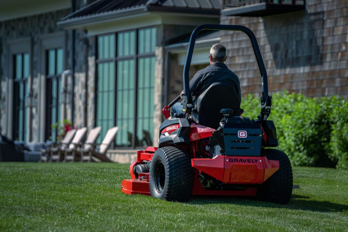 A person operates a red Gravely lawn mower on a grassy lawn near a large house with stone and siding exterior.
