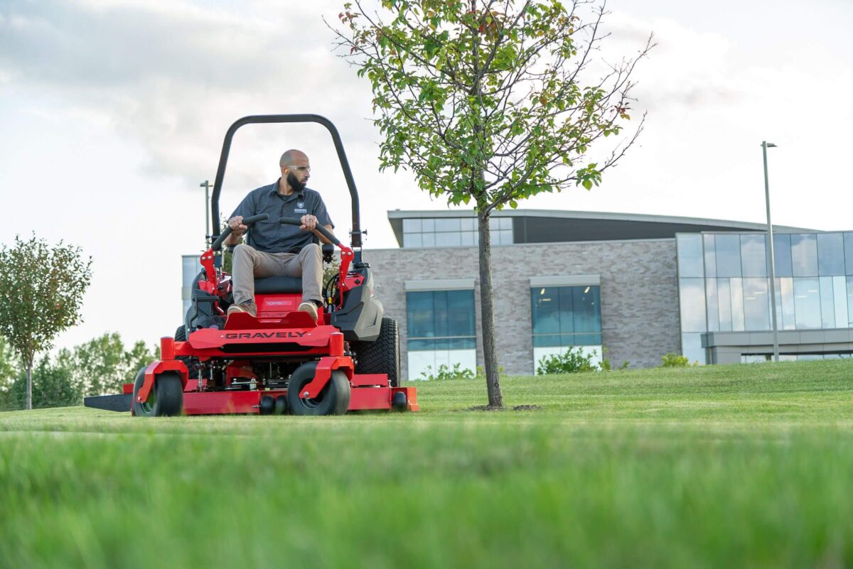 A person is riding a red Gravely lawn mower on green grass near a small tree, in front of a modern building under a partly cloudy sky.
