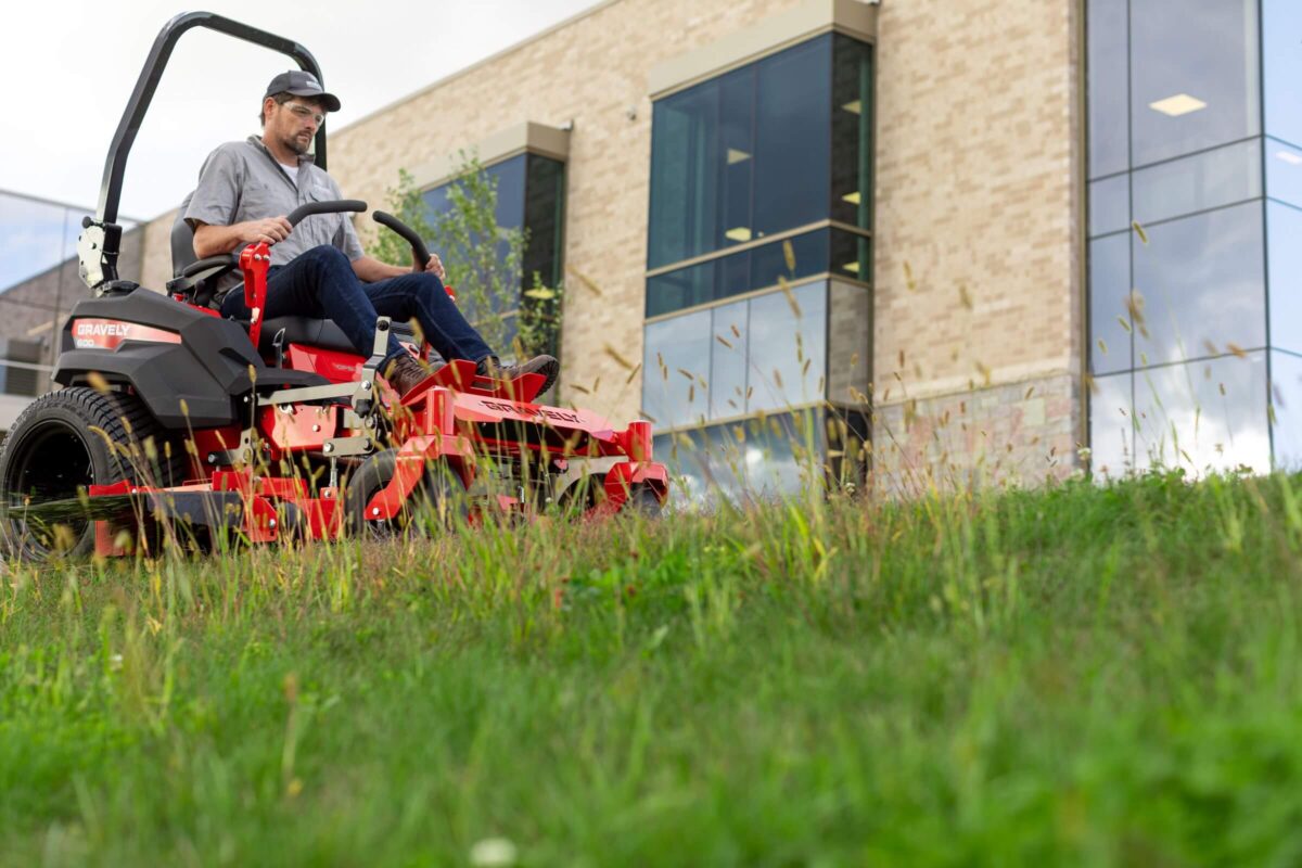 A person in a cap operates a red riding lawnmower on a grassy slope in front of a modern brick building.
