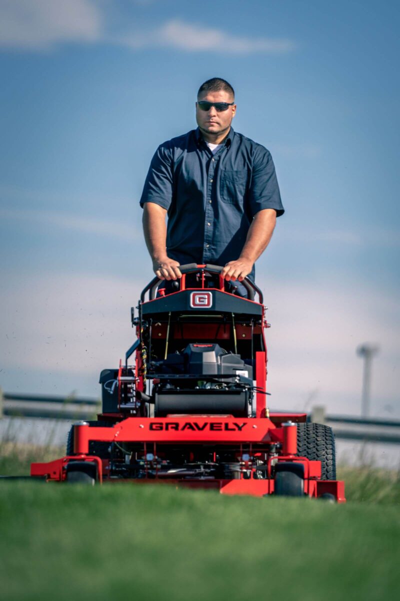 A person in sunglasses operates a red Gravely lawn mower on a grassy area under a clear blue sky.
