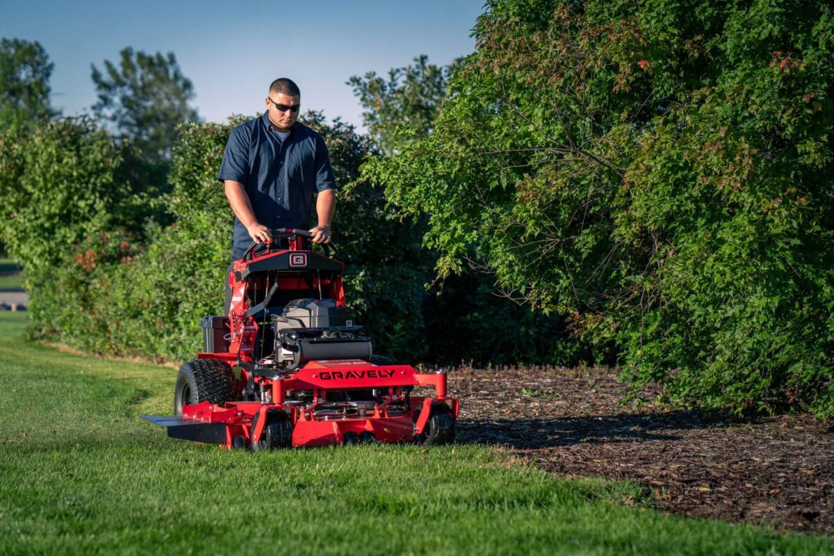 Person operating a red Gravely lawn mower on green grass beside a shrubbery.