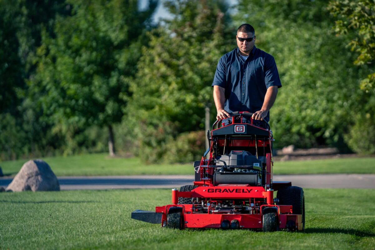 Person mowing a lawn with a red Gravely lawnmower on a sunny day, wearing sunglasses and a blue shirt, surrounded by greenery.